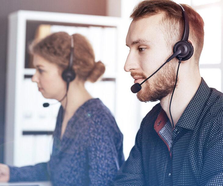 Zwei Personen mit Headsets sitzen an einem Tisch, konzentriert auf ihre Arbeit in einem modernen Büro.