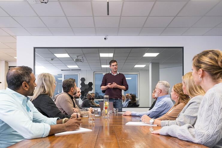 Ein Mann steht an einem Tisch und spricht zu einer Gruppe von sieben Personen in einem Konferenzraum.