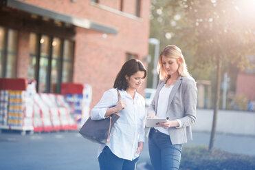 Zwei Frauen betrachten gemeinsam ein Tablet vor einem Gebäude, umgeben von Einkaufskörben im Hintergrund.