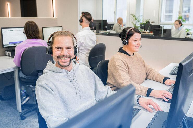 Lächelnder Mitarbeiter mit Headset an einem Schreibtisch, während eine Kollegin am Computer tippt. Offene Büroumgebung.