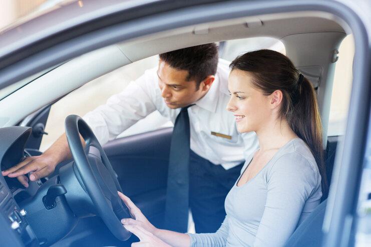 Ein Mann erklärt einer Frau in einem Auto die Bedienung des Armaturenbretts. Beide wirken aufmerksam und freundlich.