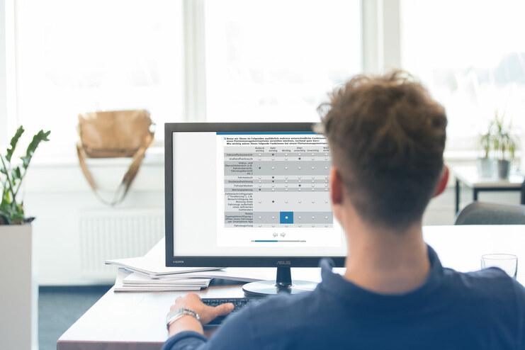 Eine Person sitzt an einem Schreibtisch und arbeitet an einem Computer mit einer Übersichtstabelle.