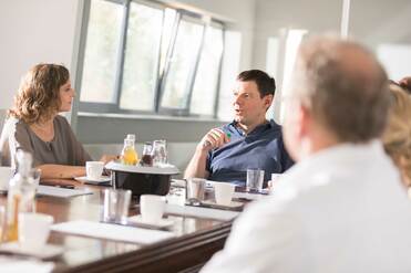 Zwei Personen im Gespräch an einem Tisch, umgeben von Getränken und Notizmaterialien in einem hellen Raum.
